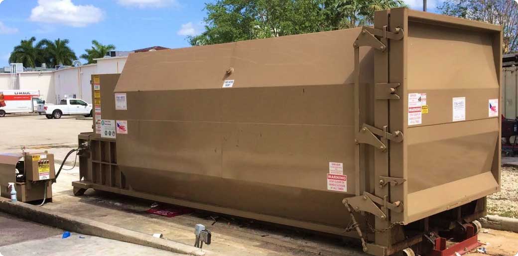 Large brown industrial trash compactor sits on a concrete pad outdoors, surrounded by warning labels. Trees and vehicles are visible in the background under a blue sky with scattered clouds.