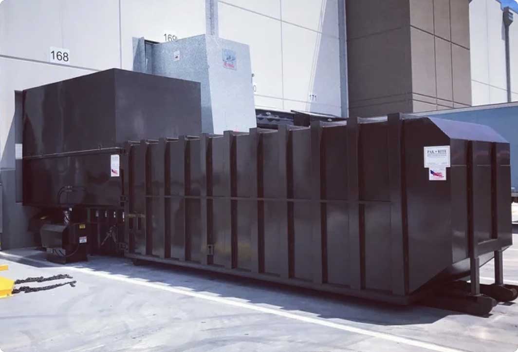 A large industrial trash compactor sits outside a commercial building near a loading dock, positioned against a white wall with numbered doors.