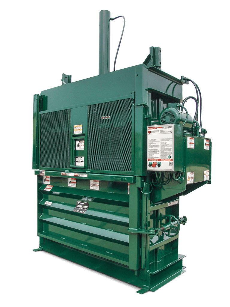 A large, industrial green baler machine with metal panels, warning labels, a control box, and hydraulic components, used for compacting cardboard or recyclable materials.